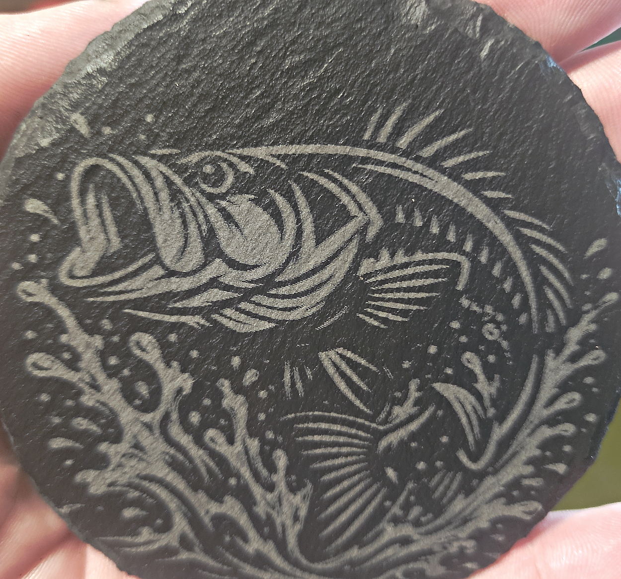 A round slate coaster held in a hand, engraved with a leaping bass surrounded by water and splashes, every line cut clean.