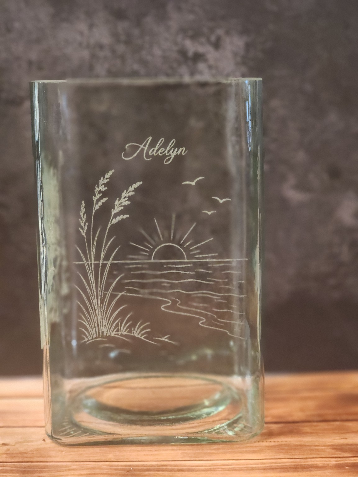 A glass vase engraved with a name in script above a beach sunset scene.
